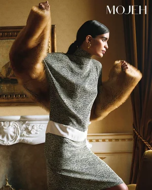 Model Bianca Martinello wearing a grey mock-neck dress and plush fur stole posing in an opulent room with a marble fireplace.