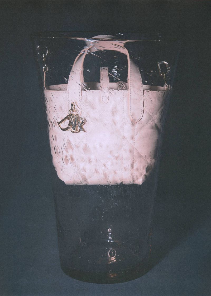 Light pink textured Dior handbag encased in a clear acrylic container with a silver logo charm, highlighting fashion and artistic display.