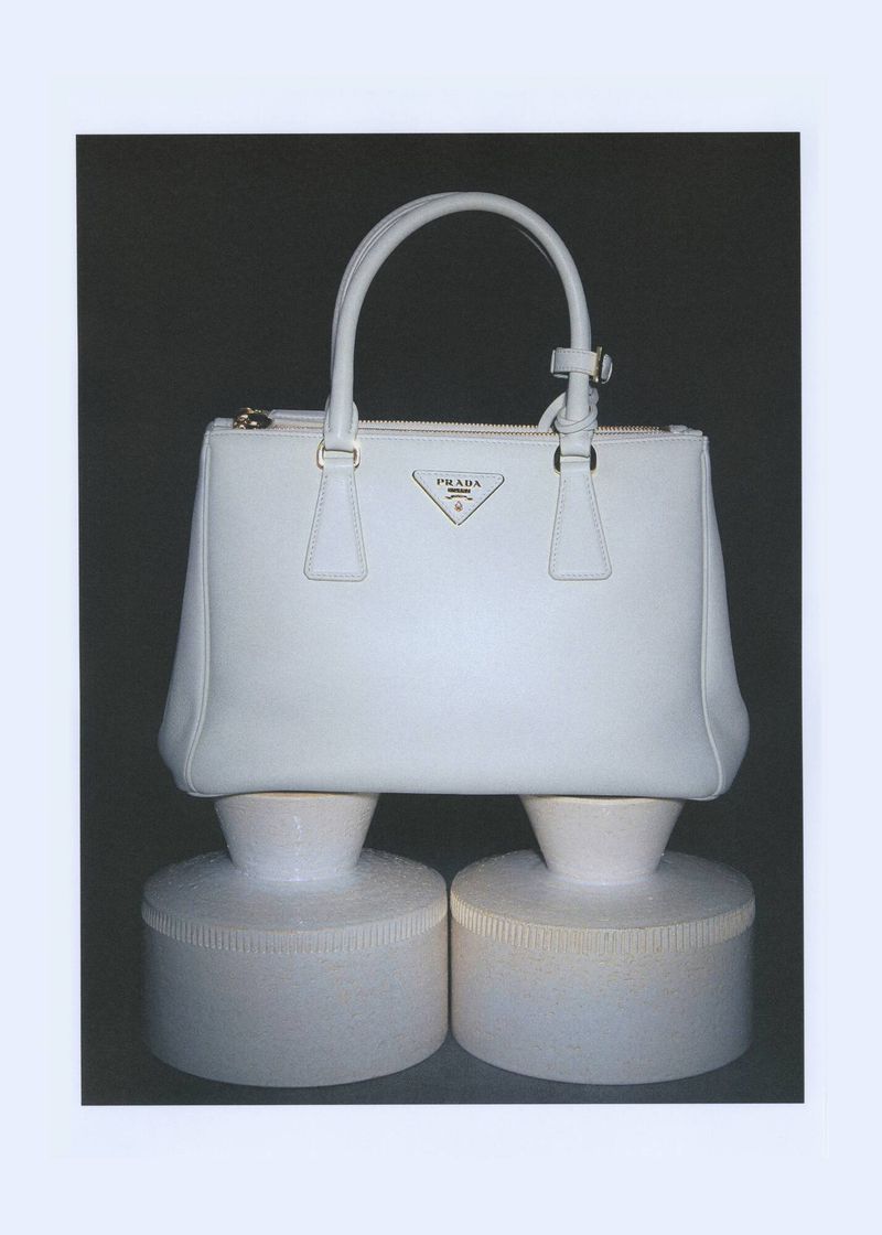 Studio shot of a white Prada handbag with handles, zippers, and a triangle logo, displayed on two cylindrical white display pedestals.