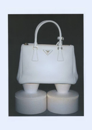 Studio shot of a white Prada handbag with handles, zippers, and a triangle logo, displayed on two cylindrical white display pedestals.
