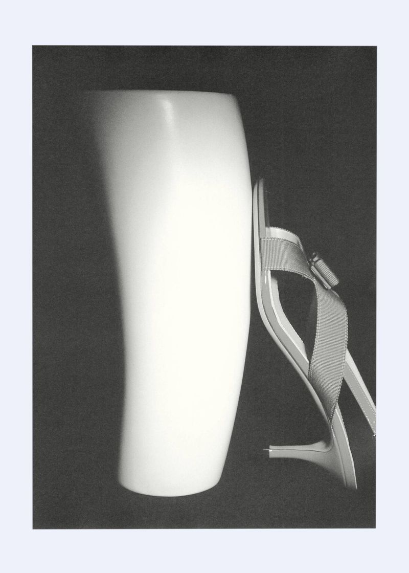 Still life photograph featuring a white ceramic vase next to a single high heel shoe on a dark background; the style is minimalist.