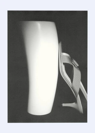 Still life photograph featuring a white ceramic vase next to a single high heel shoe on a dark background; the style is minimalist.