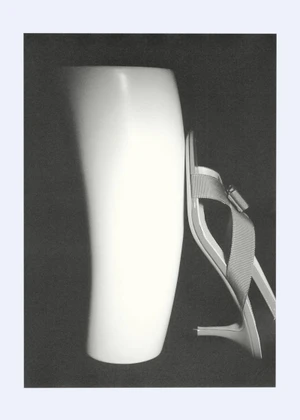 Monochrome still life of a strappy kitten heel sandal leaning against a smooth white curved sculptural form on a dark backdrop.