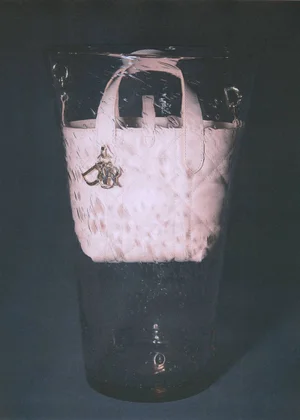 Pale pink quilted mini handbag with charm pendant suspended inside a tall clear glass vase against a dark background.