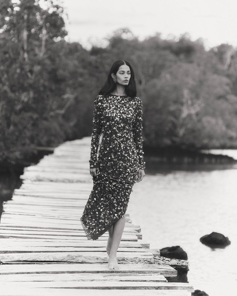 A striking black and white fashion shot featuring a lone woman in a sequined dress walking barefoot on a rustic wooden boardwalk.