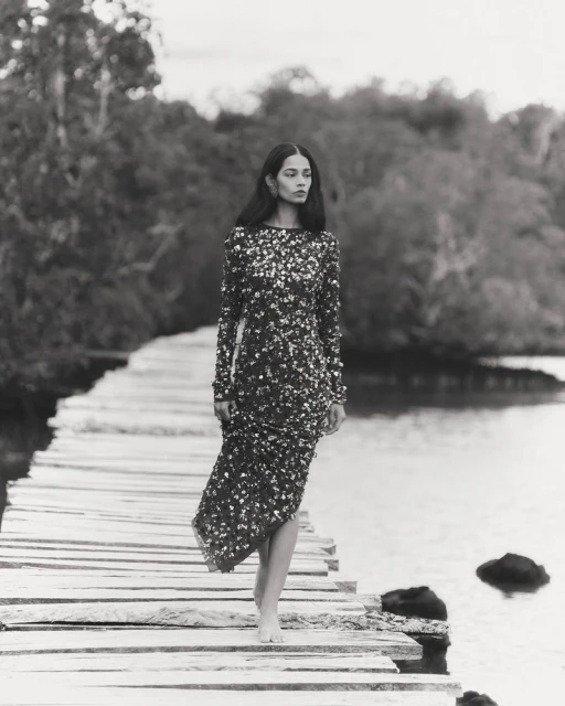 A striking black and white fashion shot featuring a lone woman in a sequined dress walking barefoot on a rustic wooden boardwalk.