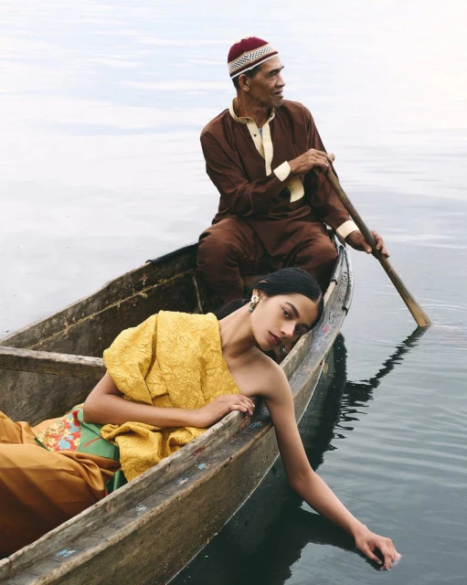 Older man rows a wooden boat with a young woman in a yellow dress; the water is calm, reflecting the sky; the scene has a serene mood.