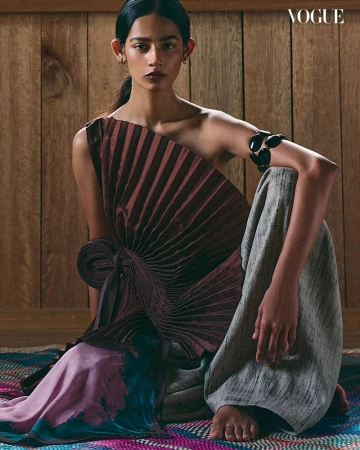 An adult female model is seated on a patterned rug against a wood panel wall, wearing a pleated brown top, wide grey pants, and Vogue logo.
