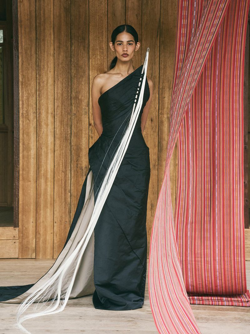 Elegant woman poses in a black one-shoulder gown with white strands accenting it, against a wooden wall with fabric draping.
