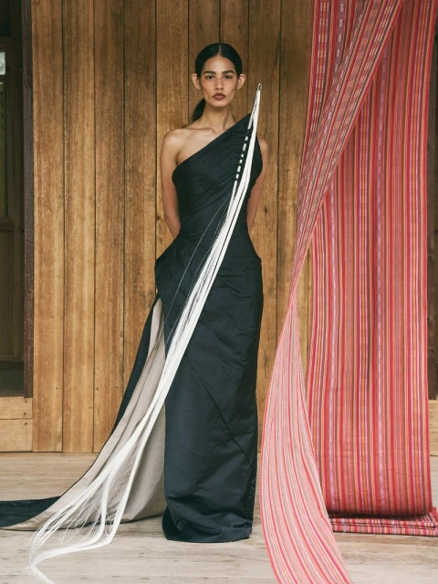 Elegant woman poses in a black one-shoulder gown with white strands accenting it, against a wooden wall with fabric draping.