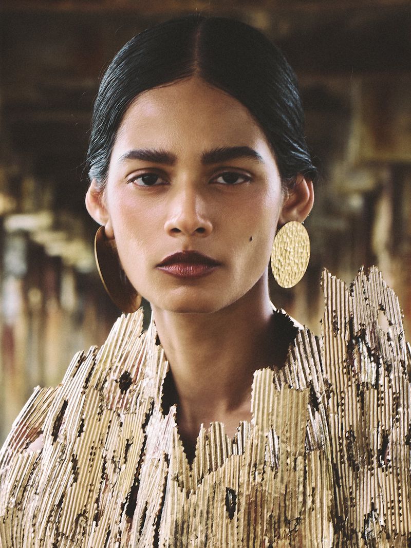 Portrait of a woman with dark hair wearing large gold earrings and a textured gold top against a blurred background.