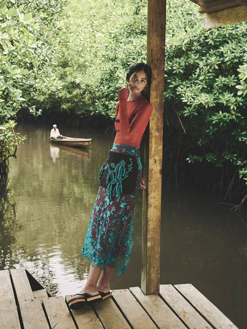 A fashionable woman poses on a dock with a waterway, wearing a red shirt and a flowing teal skirt, with a small boat on the water.