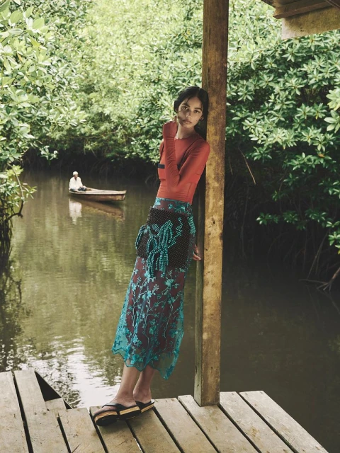 A fashionable woman poses on a dock with a waterway, wearing a red shirt and a flowing teal skirt, with a small boat on the water.