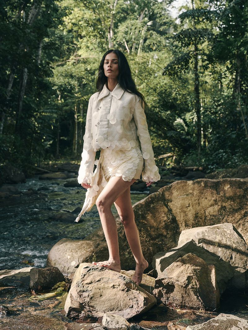 Fashionable woman in a light-colored two-piece outfit stands on rocks by a stream surrounded by a lush green forest.