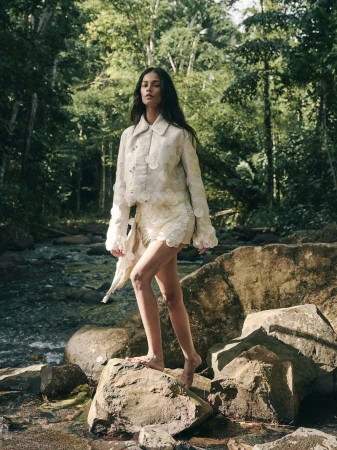 Fashionable woman in a light-colored two-piece outfit stands on rocks by a stream surrounded by a lush green forest.