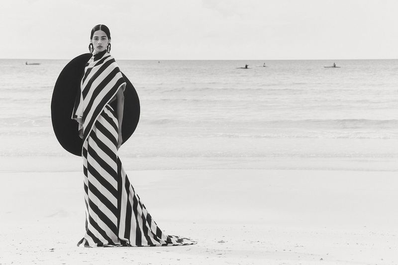 Fashion model in a black and white striped dress and large black disc-shaped hat posing on a beach near the ocean with paddle boarders.