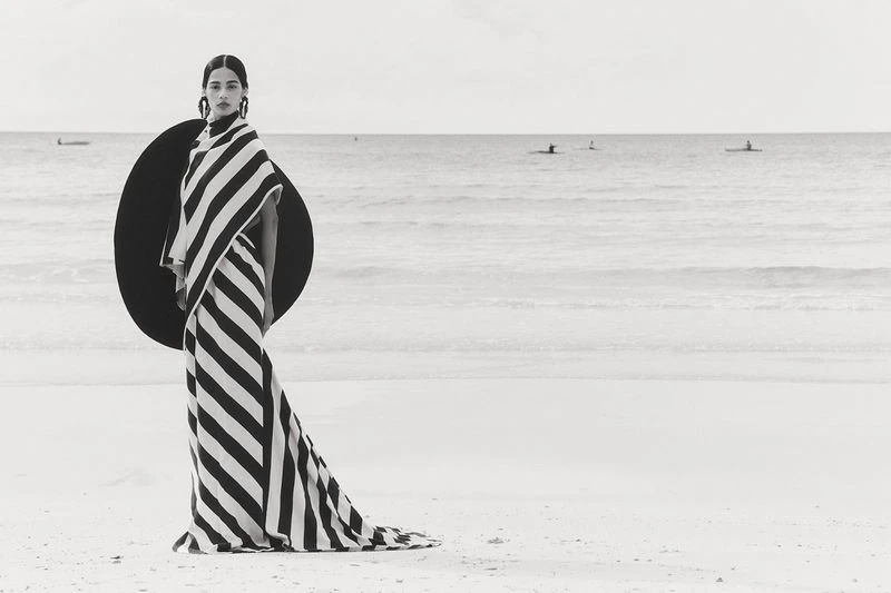 Fashion model in a black and white striped dress and large black disc-shaped hat posing on a beach near the ocean with paddle boarders.