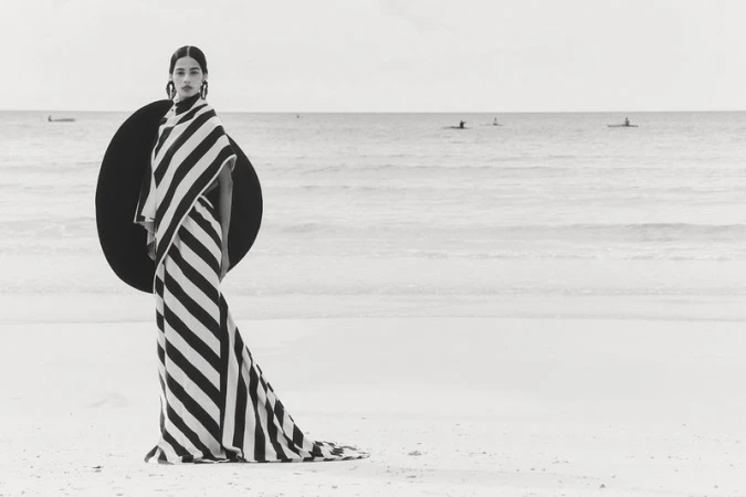 Fashion model in a black and white striped dress and large black disc-shaped hat posing on a beach near the ocean with paddle boarders.