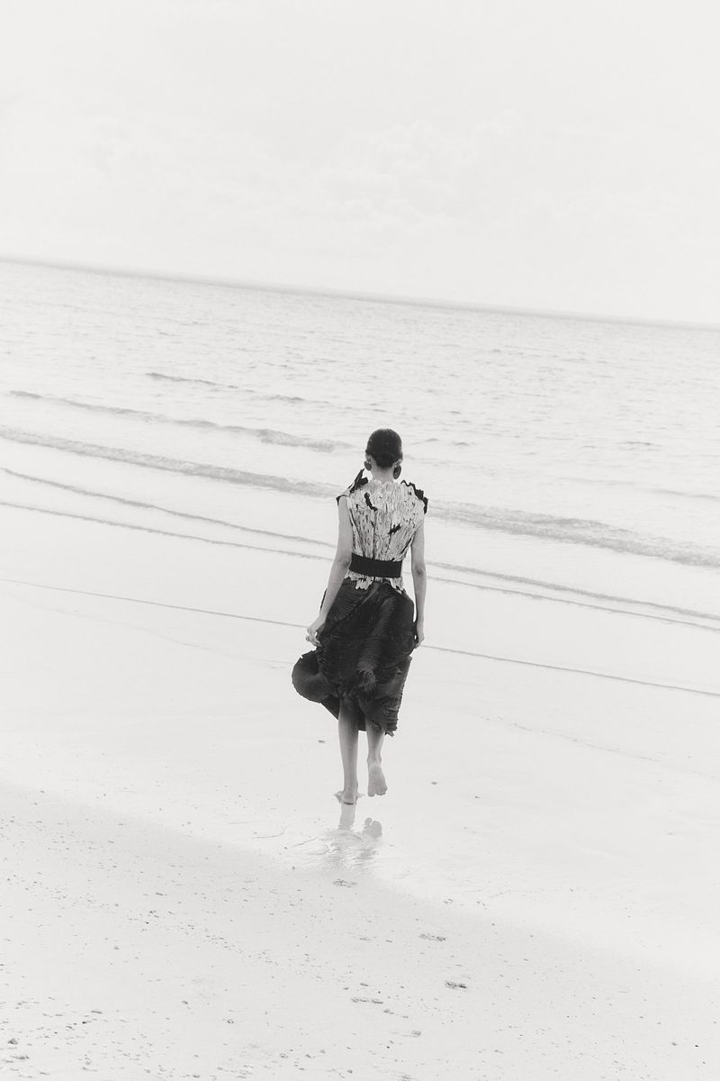 A woman walks barefoot on a sandy beach into the ocean. She wears a black and white dress and holds a hat. Ocean meets the sky.