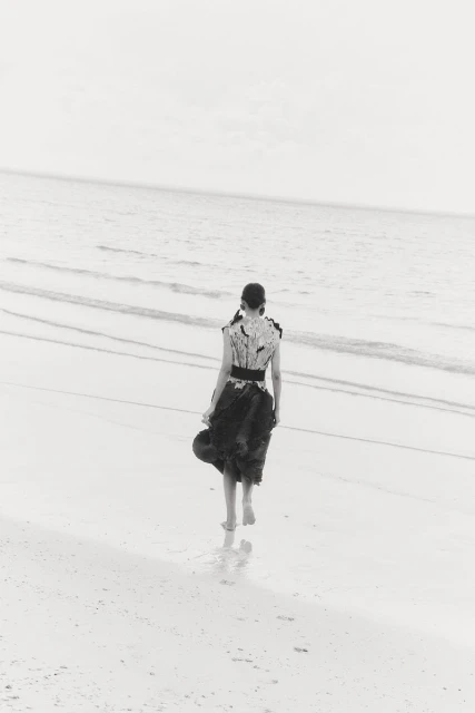 A woman walks barefoot on a sandy beach into the ocean. She wears a black and white dress and holds a hat. Ocean meets the sky.