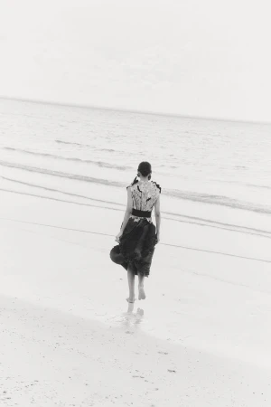 A woman walks barefoot on a sandy beach into the ocean. She wears a black and white dress and holds a hat. Ocean meets the sky.