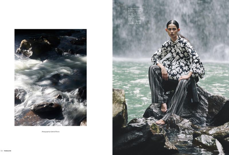 Fashion editorial shot of a woman in a floral top and striped pants sitting on rocks near a waterfall in an outdoor setting.
