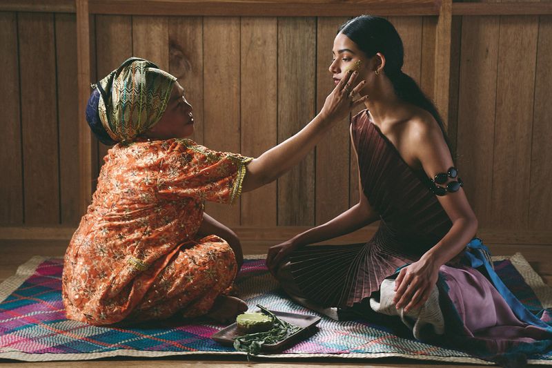 Two women indoors, one applying a yellow mask to the other's face. The scene is naturally lit, with wooden walls and a woven mat.