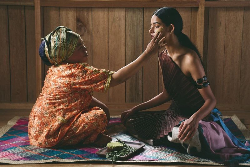 Two women indoors, one applying a yellow mask to the other's face. The scene is naturally lit, with wooden walls and a woven mat.