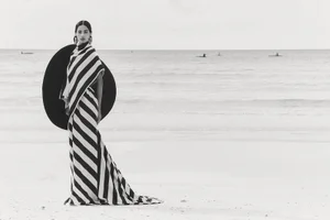 Model standing on a sandy beach wearing a striped gown and carrying a large circular black prop