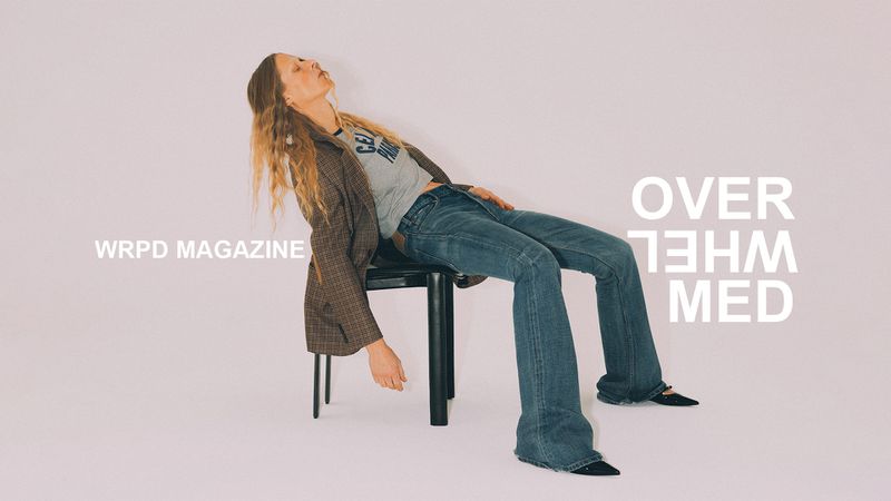 Woman in blazer and jeans poses on black stool against a pale pink backdrop with magazine text, blending fashion and editorial elements.
