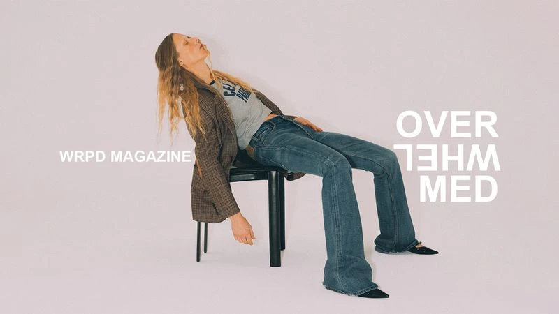 Model leaning back on a black chair wearing a plaid blazer, grey t-shirt, and blue jeans against a light background