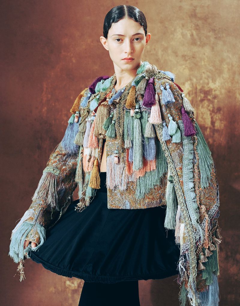 A fashionable woman in a tassel jacket poses against a textured brown backdrop, showcasing a unique and eye-catching style.