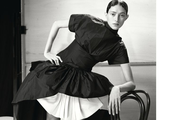Monochrome fashion portrait of an adult female model posing in a stylish black and white dress while leaning on a black chair.