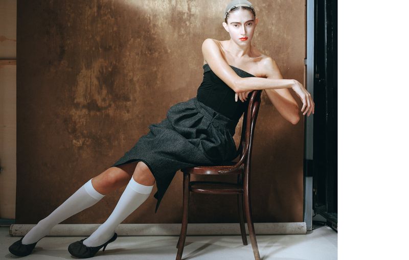 Elegant woman seated on a chair in a studio setting, wearing a strapless top, gray skirt, knee-high socks, and heels, with a neutral backdrop.