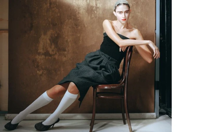 Elegant woman seated on a chair in a studio setting, wearing a strapless top, gray skirt, knee-high socks, and heels, with a neutral backdrop.