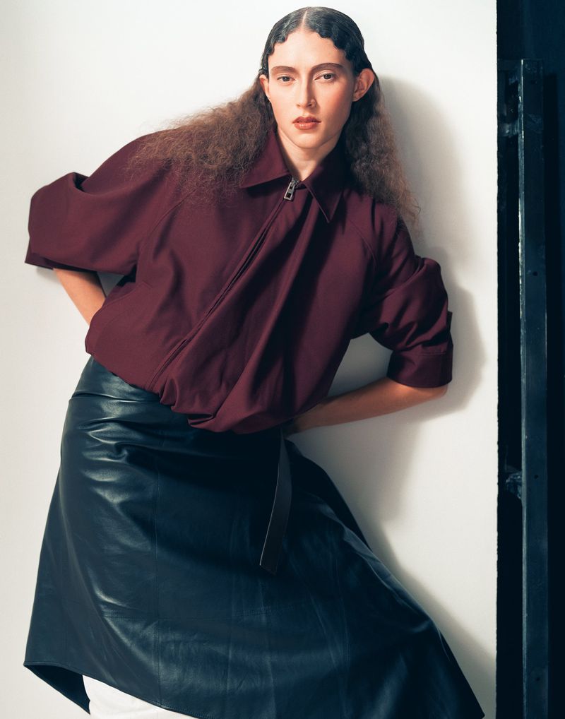 Adult female poses indoors wearing a short-sleeved maroon jacket and black skirt, complemented by her wavy brown hair.