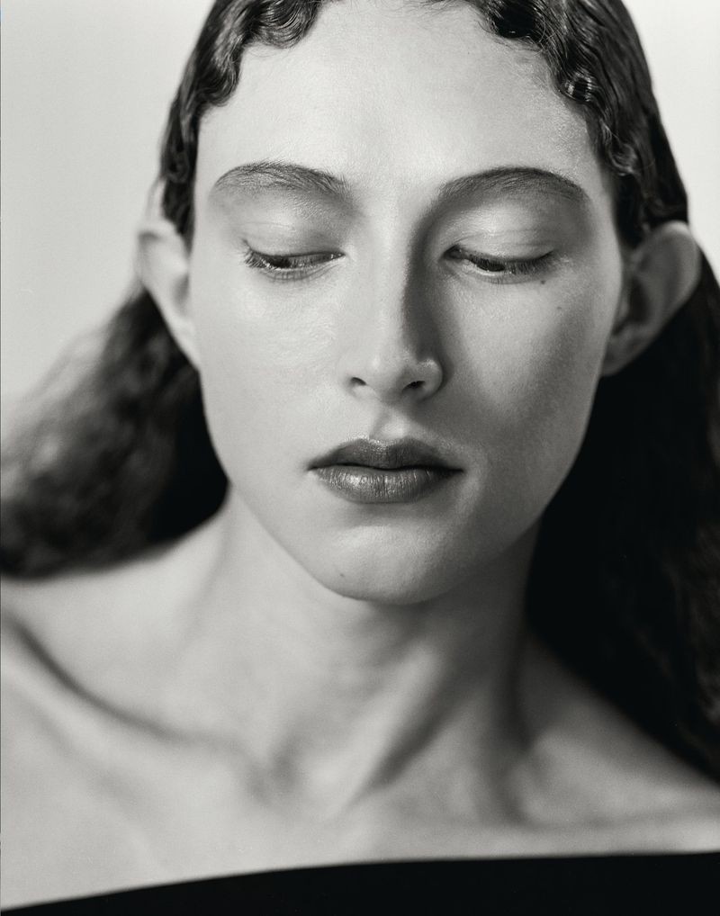 Portrait of a woman with wavy hair and dark lipstick, eyes closed, in a close-up studio shot with a neutral background and soft lighting.