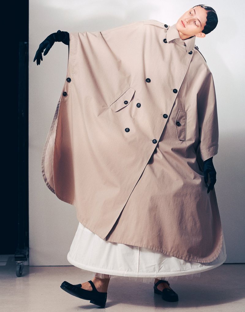 A woman wearing a beige trench coat and black gloves poses against a white wall with a dark accent, creating a high-fashion editorial shot.
