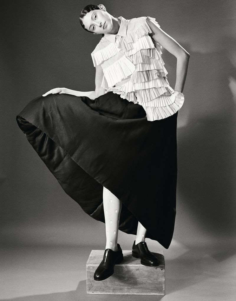 Monochrome fashion portrait of a woman in a layered white top and black skirt, standing on a wooden block in a studio setting.