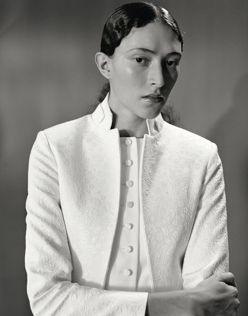 Elegant portrait of an adult female with a sleek hairstyle, wearing a white patterned jacket and top, set against a neutral backdrop.