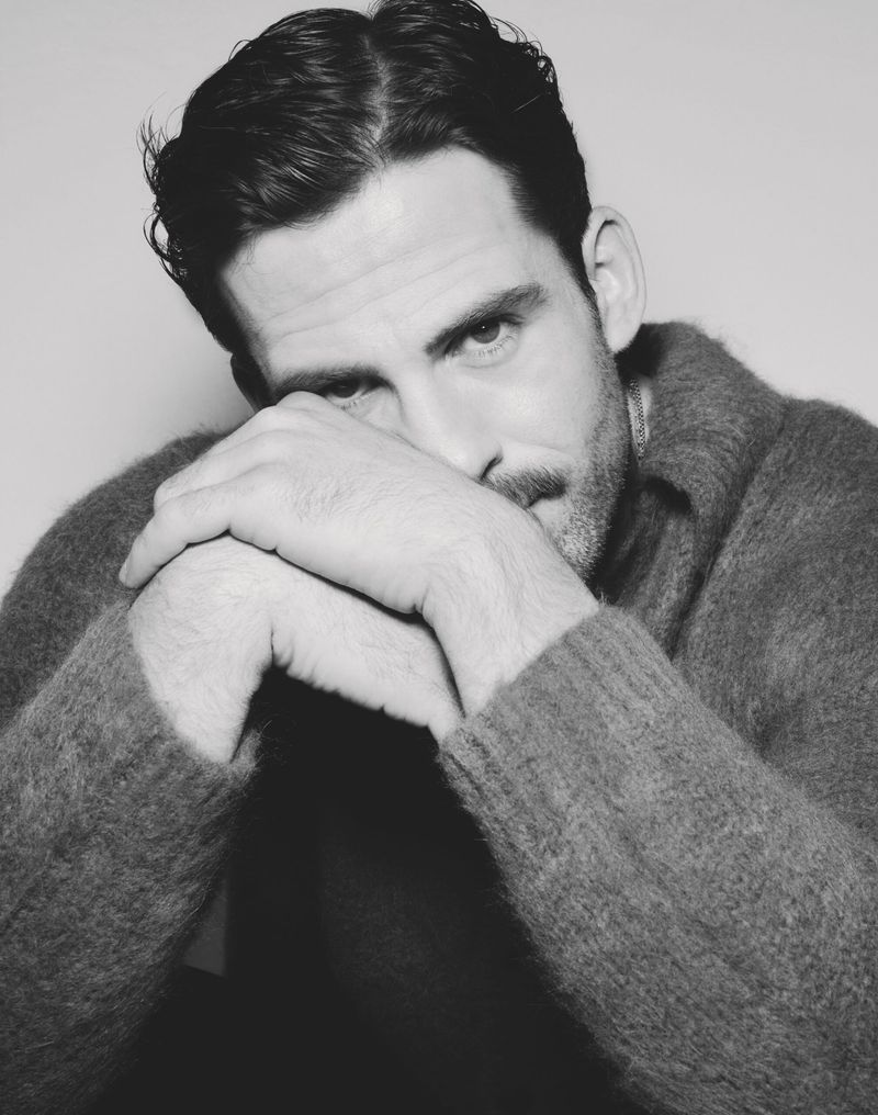 Black and white portrait of an adult man with dark hair and a mustache, wearing a cozy sweater, with his hands clasped together.