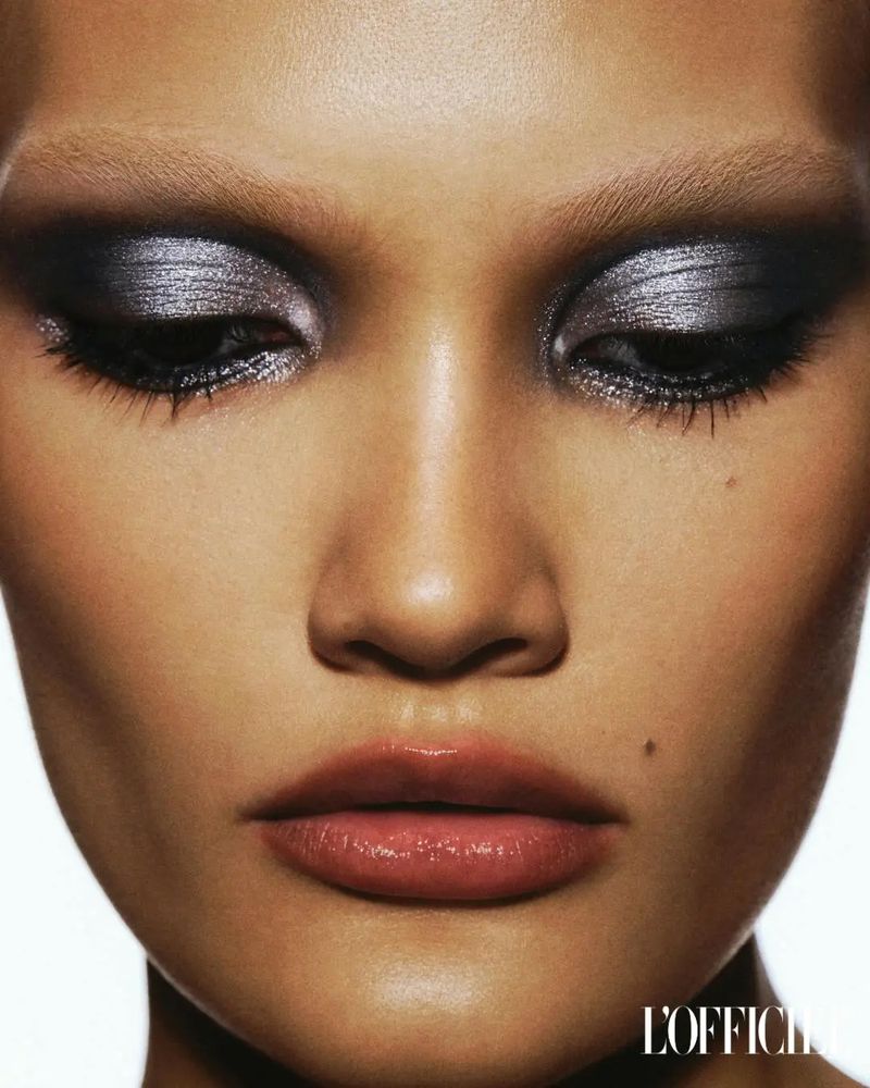 Close-up beauty shot featuring a tanned-skin adult female with silver eyeshadow and glossy lips against a clean white backdrop.