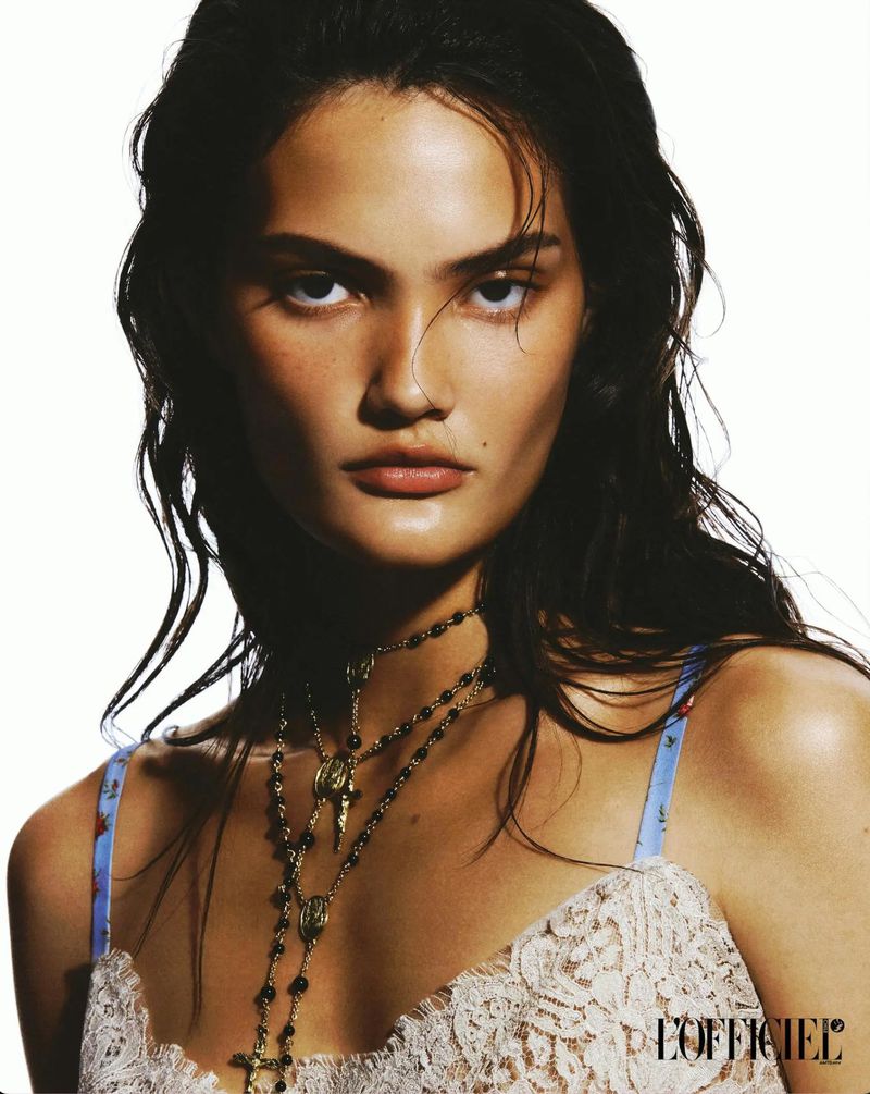 Close-up portrait of a woman with dark hair and light skin, wearing a white lace top and layered necklaces against a white background.
