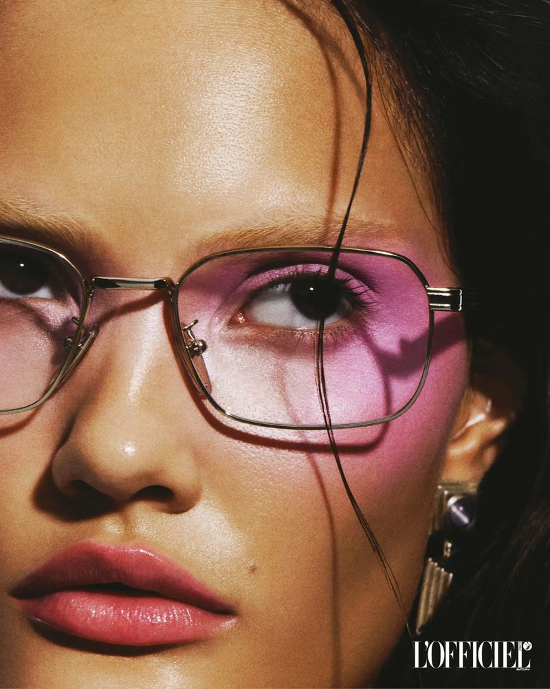 Close-up studio portrait of a woman wearing stylish eyeglasses, featuring pink lenses, and dangling earrings; minimal makeup with pink lip gloss.
