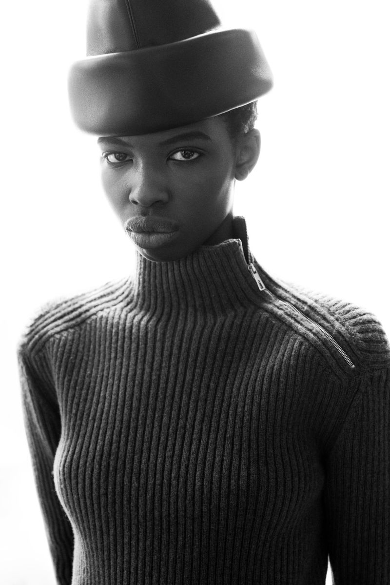 Black and white portrait of a Black female model wearing a ribbed turtleneck sweater and a dark hat, posing against a bright white background.