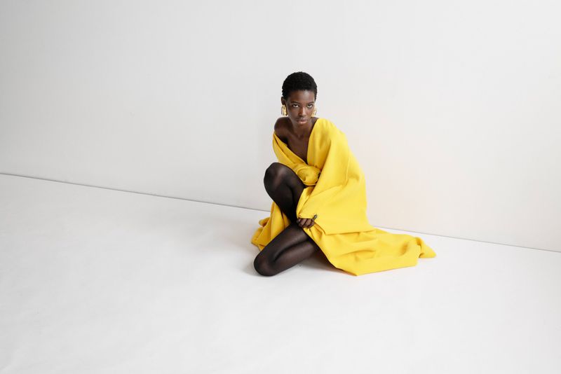Fashion portrait of an African American woman in a yellow coat and black tights, posing against a plain white backdrop in a studio setting.
