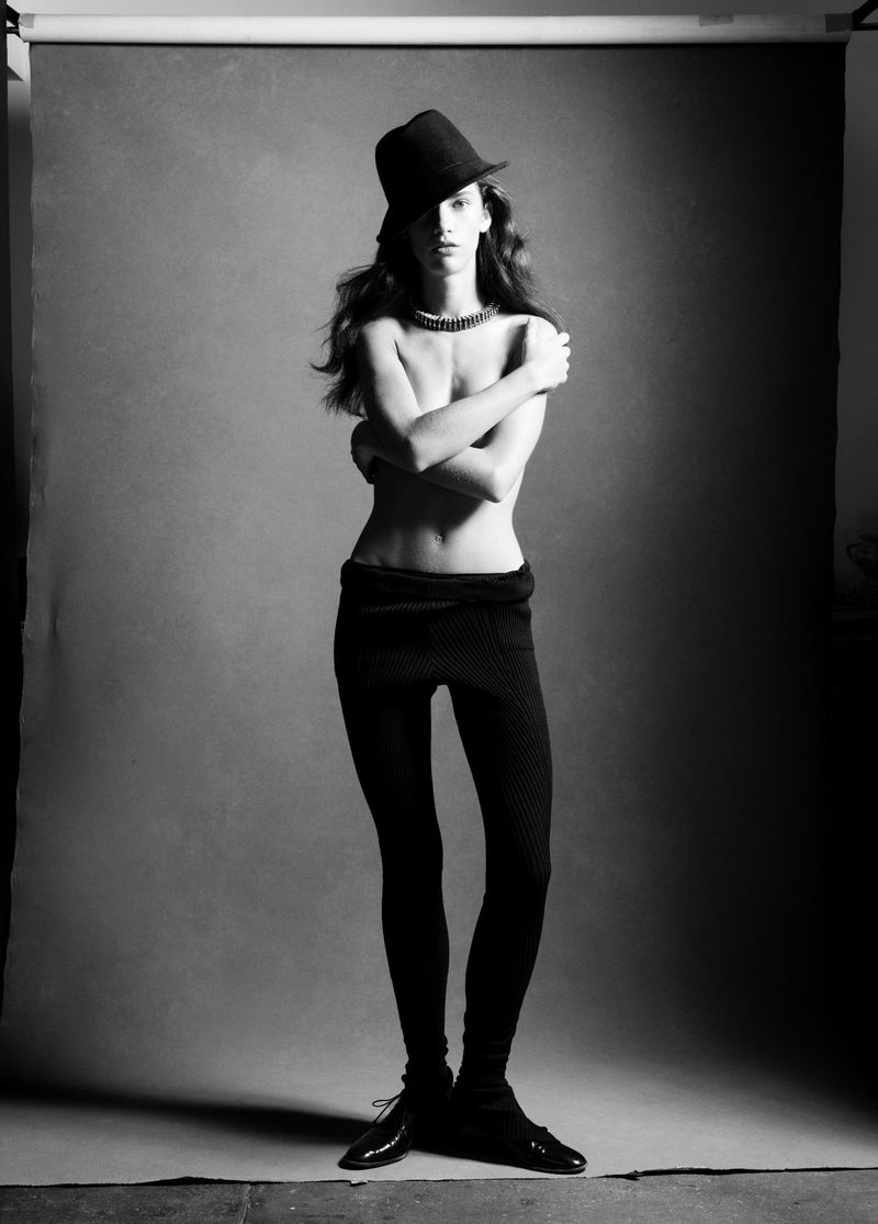 Black and white studio portrait of a topless woman with a hat, necklace, leggings, and shiny shoes against a gray backdrop.