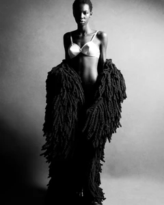 Black model posing in a white bra and an oversized black textured shaggy coat against a studio backdrop.