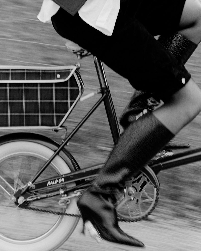 Black and white shot of a woman riding a vintage Raleigh bicycle, showcasing fashionable boots with heels and motion blur.