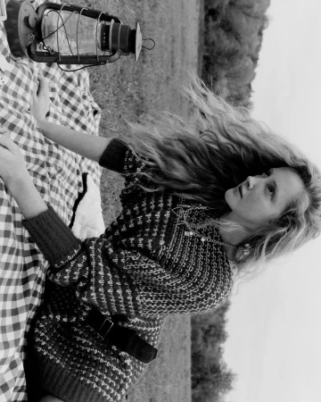Monochrome image of a young woman with long wavy hair lying on a checkered blanket with a vintage lantern, set outdoors among trees.